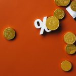Moedas douradas ao lado de um gráfico financeiro e do símbolo de porcentagem sobre fundo laranja.