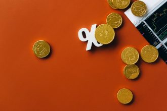 Moedas douradas ao lado de um gráfico financeiro e do símbolo de porcentagem sobre fundo laranja.