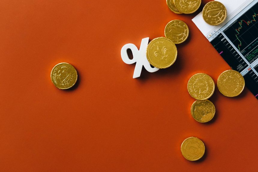 Moedas douradas ao lado de um gráfico financeiro e do símbolo de porcentagem sobre fundo laranja.