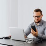 Trabalhador consulta o celular enquanto usa um laptop em uma mesa de escritório