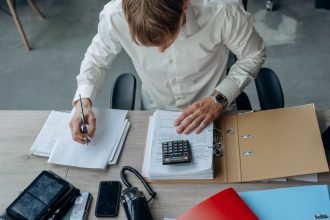 Homem analisa documentos financeiros com uma calculadora sobre a mesa em um escritório.