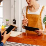 Duas pessoas interagem durante um atendimento por celular em um café; uma delas gesticula enquanto a outra toca a tela do aparelho.