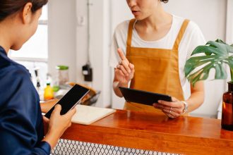 Duas pessoas interagem durante um atendimento por celular em um café; uma delas gesticula enquanto a outra toca a tela do aparelho.