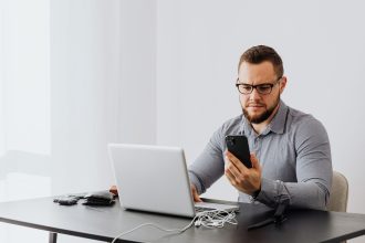 Trabalhador consulta o celular enquanto usa um laptop em uma mesa de escritório