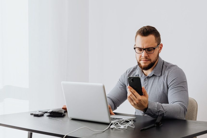 Trabalhador consulta o celular enquanto usa um laptop em uma mesa de escritório