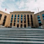 Vista externa do edifício do Federal Reserve, com bandeiras dos Estados Unidos e escadaria sob céu nublado.