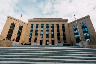 Vista externa do edifício do Federal Reserve, com bandeiras dos Estados Unidos e escadaria sob céu nublado.